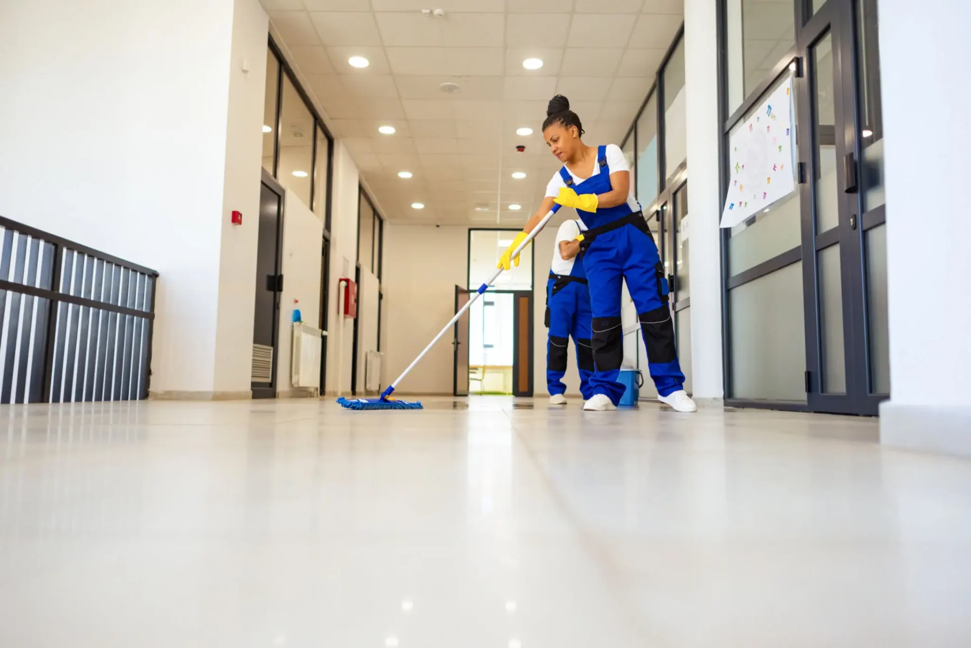 Two professional cleaners wearing blue overalls and yellow gloves mopping the floor of a modern office hallway.