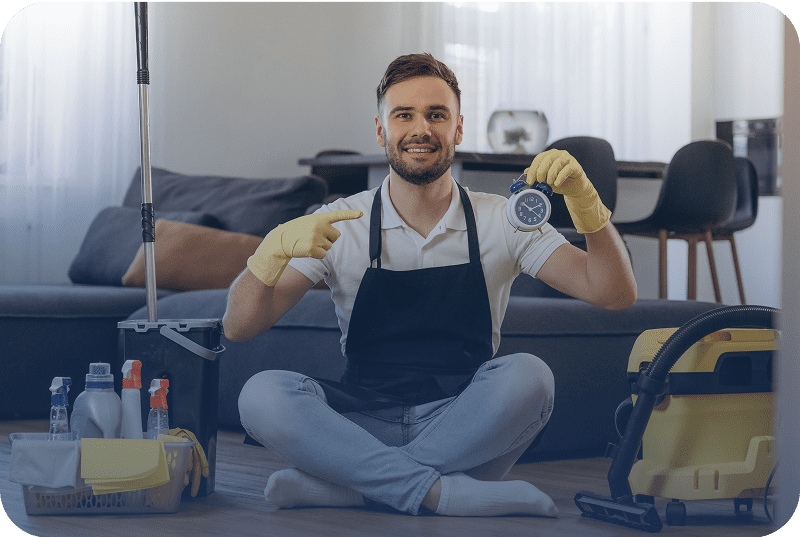 Young man in apron holding cleaning supplies and smiling.