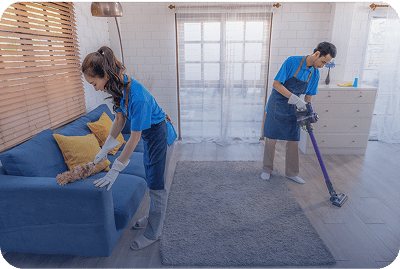 Two women cleaning a living room, one dusting a sofa, the other vacuuming the carpet.